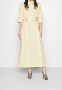 Kvinna iförd en ljusgul, lång knapp-up klänning med halvlånga ärmar och ett bälte, i kombination med svartvitt zebramönstrade klackskor.