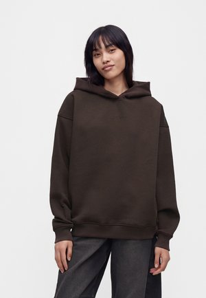 Ung kvinde med lige mørkt hår iført en mørkebrun oversized hættetrøje og mørke bukser mod en ensfarvet lys baggrund.