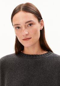 Junge Frau mit langen braunen Haaren, leichten Sommersprossen und natürlichem Makeup, die einen dunkelgrauen Strickpullover vor einem schlichten weißen Hintergrund trägt.