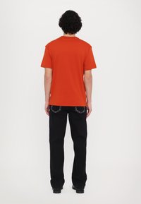 Orange short-sleeve t-shirt with a round neckline, worn with black jeans featuring contrasting stitching details, viewed from the back.