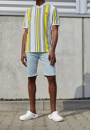 Keltainen ja valkoinen raidallinen lyhythihainen polo-paita, jossa on mustia viivoja, yhdistettynä vaaleansinisiin shortseihin ja valkoisiin puukenkään. Yksinkertainen muotoilu ja rento istuvuus.