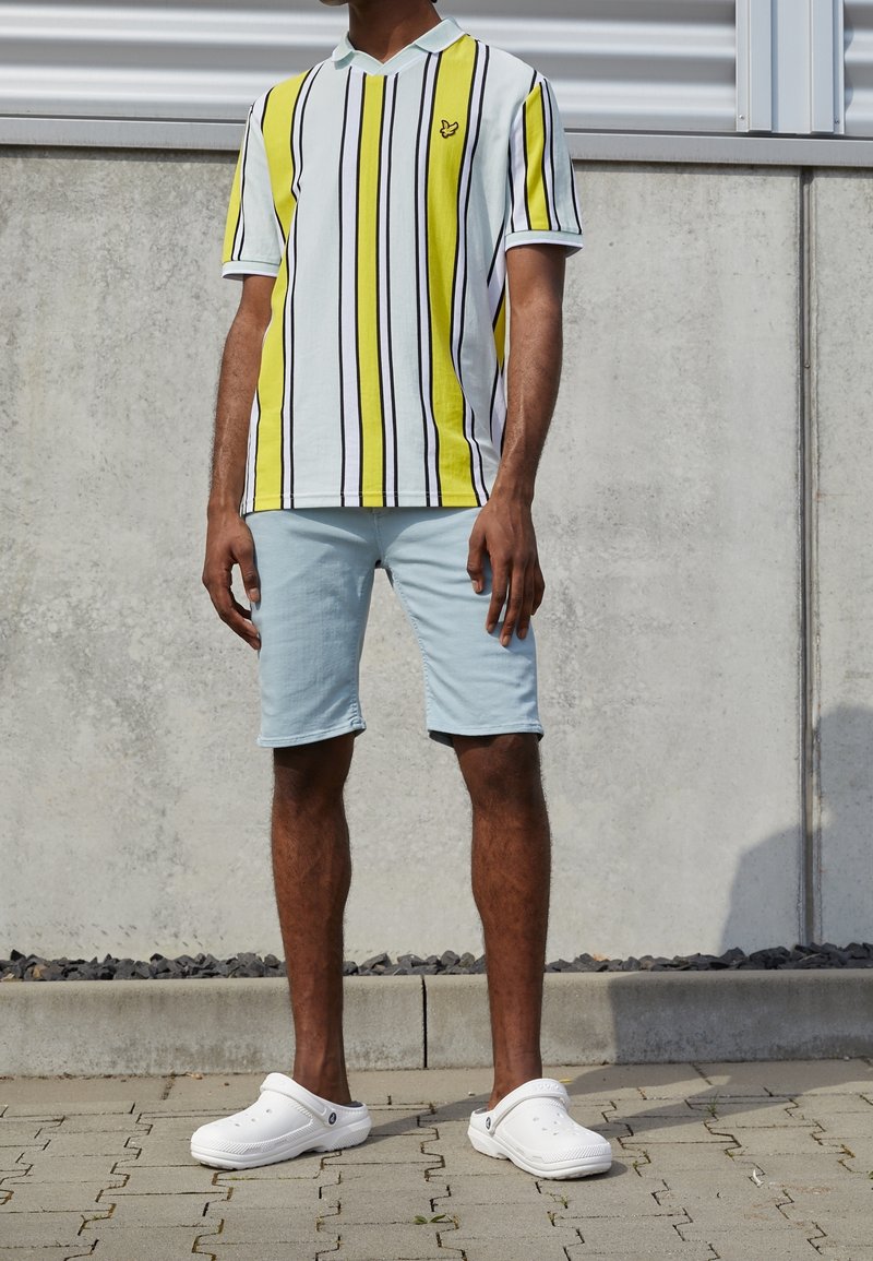 Polo shirt à rayures jaunes et blanches à manches courtes avec des lignes noires, associé à un short bleu clair et des sabots blancs. Design simple et coupe décontractée.