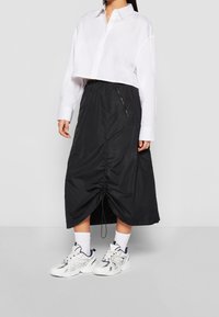 Chemise blanche coupée avec une jupe midi en nylon noir dotée d'un ourlet à cordon de serrage et de poches latérales zippées. Portée avec des baskets blanches.