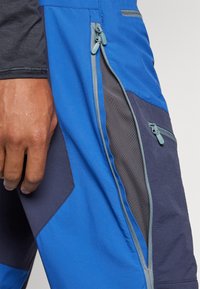 Blue and navy outdoor pants featuring multiple zippered pockets and contrasting fabric panels in breathable material. Seam detail along edges.