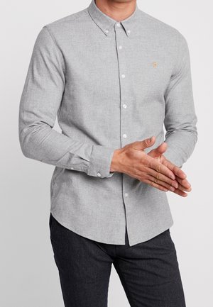 Man draagt een lichtgrijs nauwsluitend overhemd met lange mouwen en donkere broek, met de handen samengevouwen voor zich.