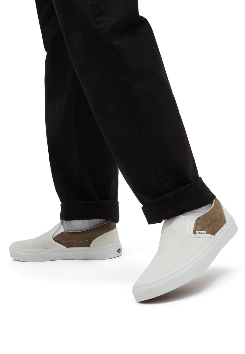 Persona che indossa sneakers slip-on bianche e marroni con calze bianche e pantaloni neri arrotolati, in piedi su uno sfondo bianco.