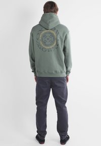 Sweat-shirt à capuche vert avec un graphique circulaire et du texte au dos, associé à un pantalon sombre et des chaussures noires. Design simple, style décontracté.