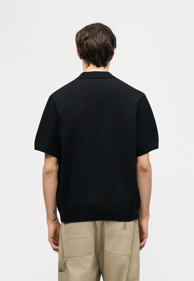 Mand med kort brunt hår iført en sort kortærmet skjorte og beige bukser, set bagfra mod en ensartet hvid baggrund.