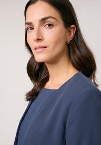 Navy blue top with a round neckline and textured fabric, featuring a smooth finish. Model has medium-length dark hair and subtle earrings.