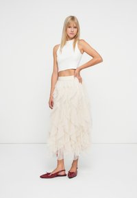 Kvinne står med hånden på hoften, iført ermeløs hvit crop top, lagdelt beige tyllskjørt og røde slingback-sko mot en enkel hvit bakgrunn.