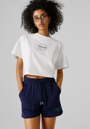 Witte cropped T-shirt met groene grafische tekst; gecombineerd met marineblauwe korte broek met elastische tailleband en zijzakken.