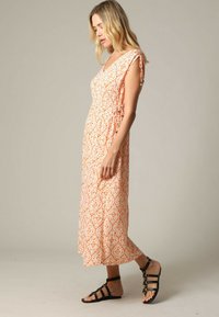 Femme portant une robe midi sans manches orange à motifs avec des liens latéraux, chaussée de sandales plates à lanières noires, se tenant devant un fond neutre.