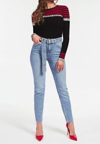 Musta ja punainen raidallinen pitkähihainen paita, jossa on logonauha, yhdistettynä vaaleansinisiin korkeavyötäröisiin farkkuihin ja punaisiin kärkitossuihin.