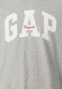 Tricou din bumbac gri, cu litere albe mari care scriu "GAP" și cuvântul "Original" scris cu cursiv roșu dedesubt. Textură moale, design casual.
