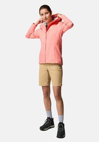 Giacca impermeabile rosa chiaro con cappuccio, chiusura con zip e polsini elastici, abbinata a pantaloni beige e scarponi da trekking neri.