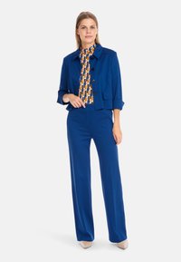Blue cropped jacket with large buttons, paired with printed high-neck top in orange, blue, and white. Wide-leg blue trousers and beige heels.