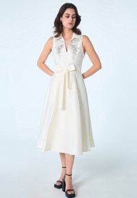 Robe blanche sans manches, longueur genoux, avec un col, broderies florales au niveau du col, et une ceinture en satin nouée à la taille.