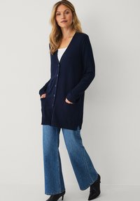Marineblauwe lange cardigan met V-hals, knoopsluiting, twee voorzakken en lange mouwen, gecombineerd met flared jeans en hakken.