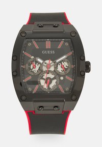 Montre noire avec accents rouges, boîtier rectangulaire, cadran squelettique, trois sous-cadrans et bracelet noir texturé. Index des heures audacieux en rouge.