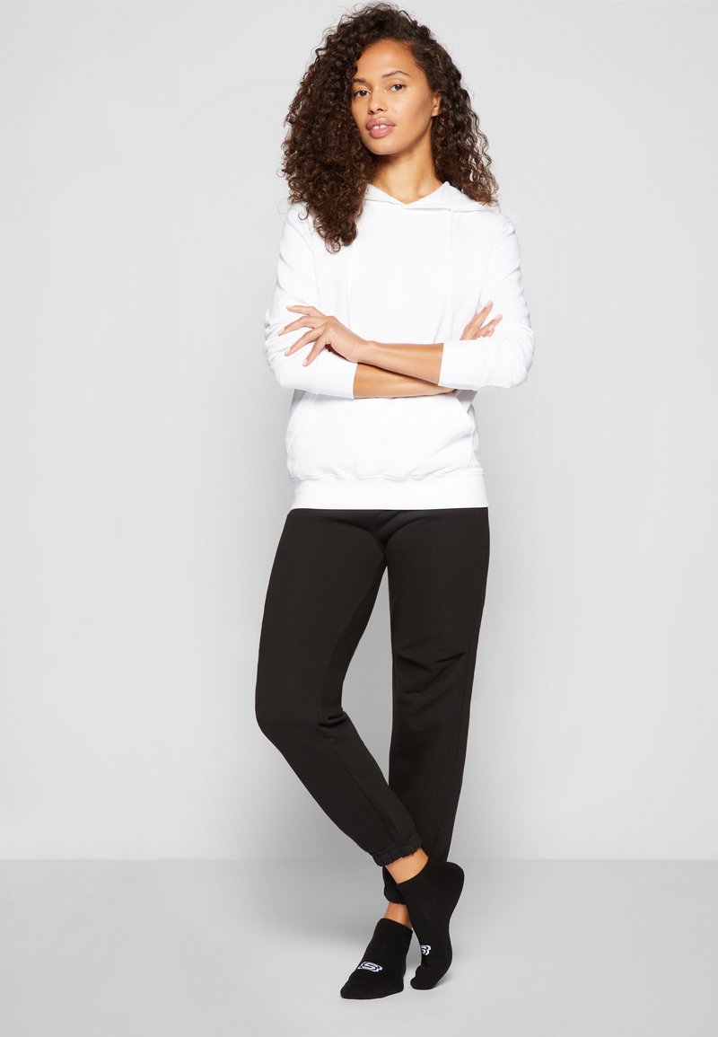 Un sweat à capuche blanc avec poche avant et cordons, associé à un pantalon de survêtement noir avec des poignets élastiques et des chaussettes noires avec logo.