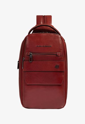 Mochila bandolera compacta de cuero rojo con bolsillo delantero con cremallera, asa superior y correa ajustable. Logo metálico de la marca en el panel frontal.