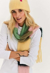 Kobieta z długimi blond włosami, w żółtej dzianinowej czapce, wielokolorowym szaliku, białej koszulce z długim rękawem, pokazująca tatuaż węża na przedramieniu.