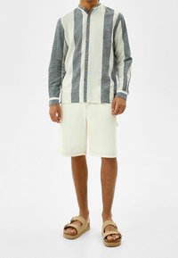 Knapp-up skjorta med marinblå och vita vertikala ränder, lättviktig tyg, mandarin-krage; tillsammans med ljusbeige shorts och sandaler.