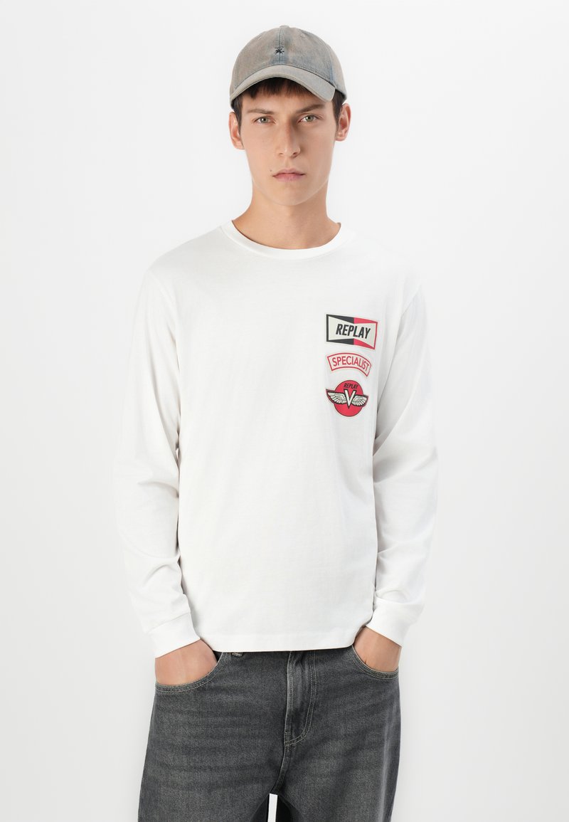 Jonge man met een grijze pet, een witte lange mouwen shirt met rode en zwarte logo's, en donkergrijze jeans, met zijn handen in zijn zakken, staand tegen een witte achtergrond.