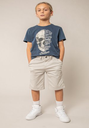 Garçon debout, les mains dans les poches, portant un t-shirt bleu marine avec un motif de crâne, un short beige, des chaussettes blanches et des baskets montantes blanches.