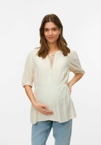 Creme farvet graviditet bluse med korte puffærmer, frontknaplukning og snoretræk i taljen, parret med lyseblå jeans.