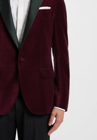 Burgunderroter Samtblazer mit einem dunkelgrünen Satin-Schalkragen, einreihiger Knopfverschluss und zwei seitlichen Taschen. Weißes Taschentuch als Akzent.