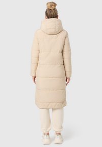 Beige pufferjack met capuchon, met geborduurde details en een lange silhouette. Gekleed met crèmekleurige joggingbroek en lichte sneakers.