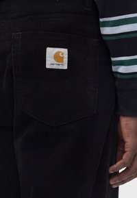 Τσέπη μαύρου παντελονιού από κορντούρα με λευκή ετικέτα της μάρκας Carhartt, δίπλα σε χέρι και ριγωτό μανίκι.