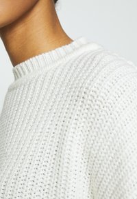 Gros plan sur le cou et l'épaule d'une personne portant un pull blanc en tricot avec un col côtelé, sur un fond uni.