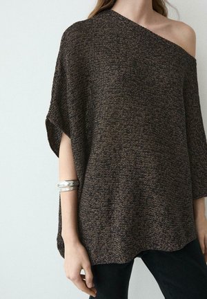 Donna che indossa una maglia lavorata a maglia marrone scuro con spalle scoperte e pantaloni neri, con un braccialetto a polsino d'argento al polso sinistro.