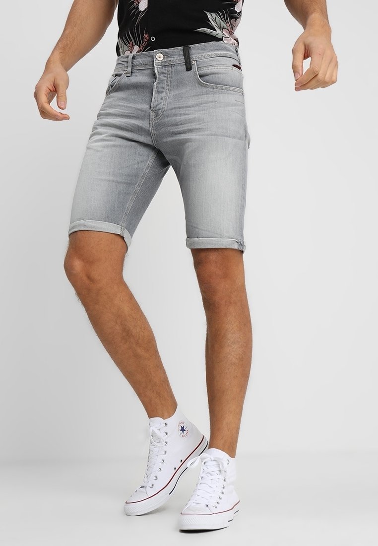 Homme portant un short en denim gris clair, une chemise noire à fleurs et des baskets blanches montantes, debout, jambes écartées, sur un fond uni.