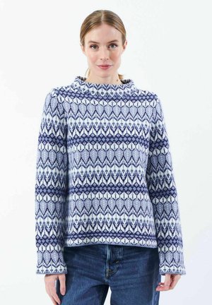 Kvinde iført en langærmet sweater med et blåt og hvidt geometrisk mønster og blå jeans, stående mod en hvid baggrund.