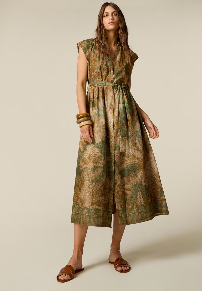 Woman wearing sleeveless brown and green patterned midi dress with buttons, bracelets on left arm, and brown flat sandals, standing against plain background.