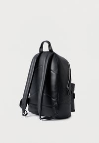 Mochila preta com acabamento em pele suave, alças de ombro acolchoadas e um bolso lateral com padrão texturizado. Fecho de correr na parte de cima.