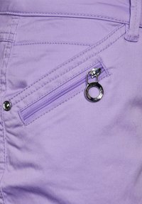 Fioletowe szorty materiałowe z kieszenią na zamek błyskawiczny z srebrnym suwakiem, kontrastujące z widocznymi przeszyciami i gładką fakturą.