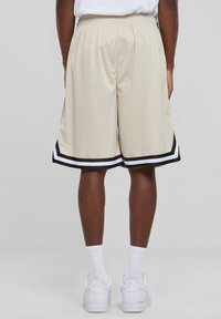 Personne portant un short de basket beige avec des rayures noir et blanc, des chaussettes blanches et des baskets blanches Nike Air, se tenant devant un fond uni.