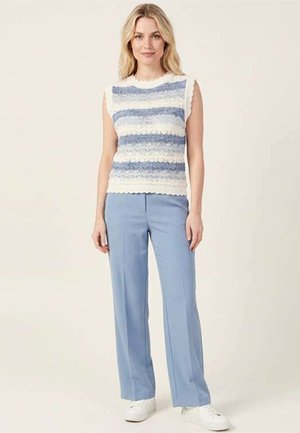 Femme debout, portant un haut tricoté sans manches rayé bleu et blanc, un pantalon large bleu clair et des baskets blanches sur un fond uni.