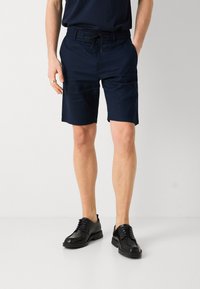 Pantaloni corti neri scuri su misura con vita elastica e cordoncino, realizzati in un tessuto liscio; abbinati a scarpe in pelle nera.