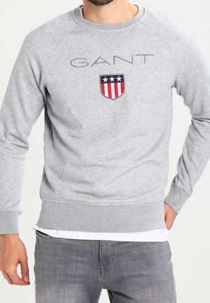 Man wearing light gray GANT sweatshirt with shield logo featuring three white stars above red and white stripes, paired with gray jeans.