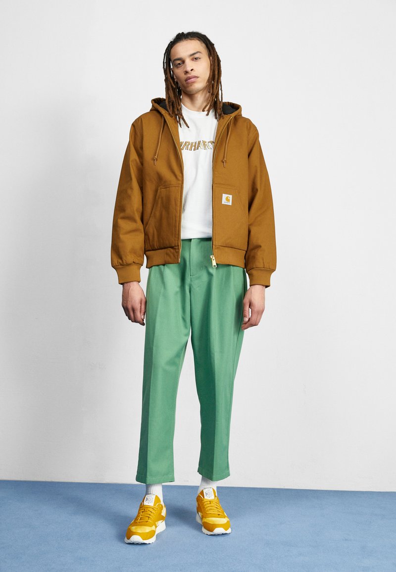 Brown zip-up hooded jacket, white shirt with logo, green trousers, and yellow sneakers with white accents, standing on blue flooring.
