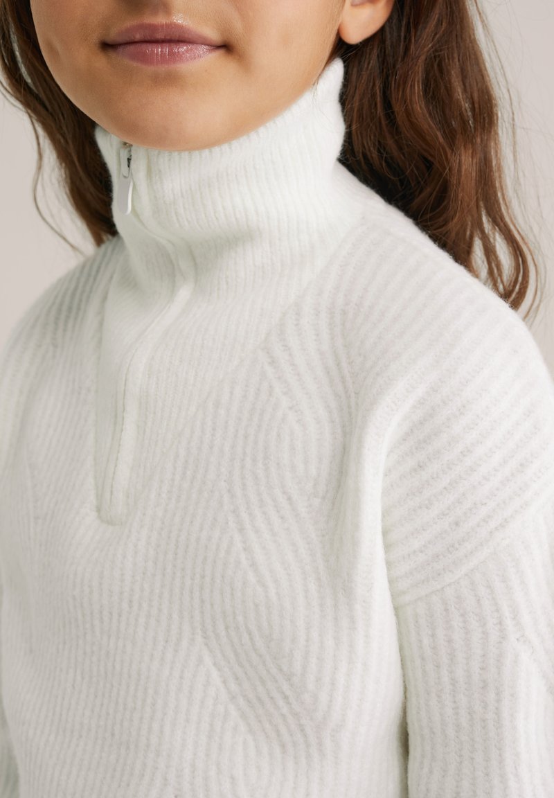 Bela zip-up jopica z visokim ovratnikom, ki ima teksturiran rebrast design in geometrijske vzorce na tkanini. Gladek, mehak material.