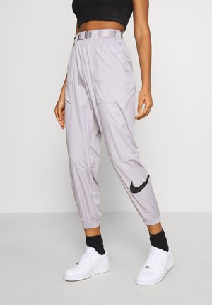 Osoba w jasnoszarych spodniach dresowych Nike, czarnym krótki topie, czarnych skarpetkach i białych butach sportowych Nike stojąca na jednolitym tle.