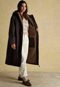 Garšs brūns mētelis ar pūkainu gaiši brūnu oderi, uzvilkts atpogāts virs rakstainā džempera un baltām biksēm. Brūnas mokasīnas pabeidz tērpu.