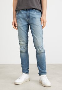 Osoba ubrana w jasnoniebieskie, przetarte dżinsy slim fit z małymi białymi plamkami farby i białe sneakersy, stojąca na gładkiej podłodze.