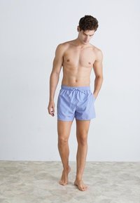 Lyseblå svømmedraktshorts laget av glatt stoff med elastisk midjebånd, snøring og en avslappet passform. Modellen står barbeint.